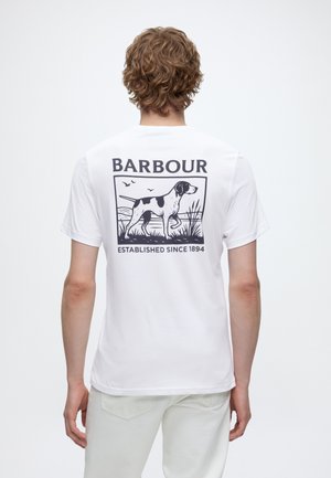 Homme aux cheveux bouclés portant un t-shirt blanc avec un graphisme de chien noir, des oiseaux, de l'herbe et le texte "BARBOUR Established Since 1894" au dos.