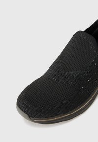 Sorte slip-on sko laget av strukturert mesh-stoff, med glitrende detaljer. Rund tå og minimal mønstring på sålen. Lett demping synlig.
