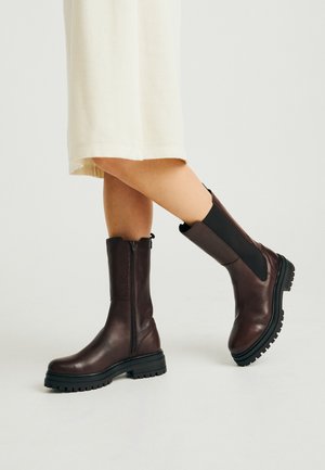 Jambes portant des bottes en cuir marron foncé mi-mollet avec des panneaux latéraux élastiques noirs et des semelles épaisses à crampons sur un fond clair.