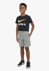 Črna majica s svetlo zelenim znakom Nike in belim besedilom, skupaj s sivimi kratkimi hlačami z belim znakom Nike. Črni supergi z belimi poudarki.