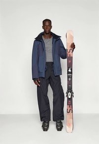 Stroje narciarski składający się z ciemnoniebieskiej kurtki z szarymi akcentami, szarej koszuli, czarnych spodni narciarskich i czarnych butów narciarskich, trzymający nartę z różowymi grafikami.