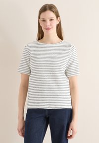 Jeune femme aux longs cheveux bruns portant un haut blanc à manches courtes avec de fines rayures horizontales noires et un jean bleu foncé, debout contre un mur beige.