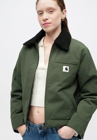 Olijfgroene bomberjack met een zwarte fleece kraag, voorrits en een klein logo patch op de borst. Gedragen over een crèmekleurige kabelbrei-top.