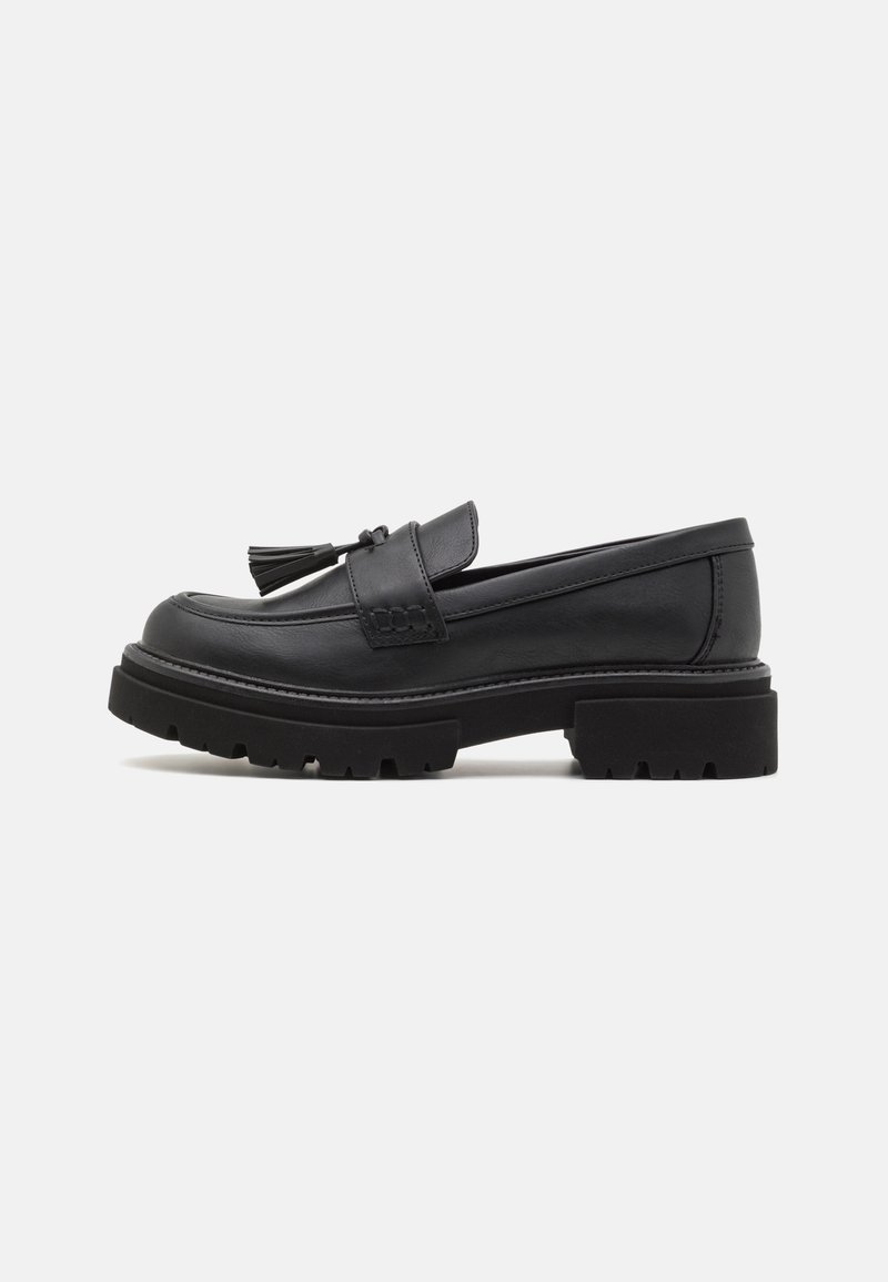 Mocassin en cuir noir avec semelle épaisse à crampons et détail pompon sur fond gris, vue de profil.