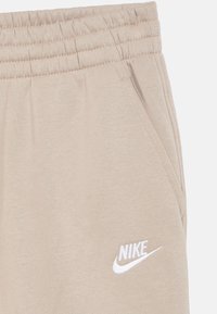Pantalon de survêtement Nike beige avec une taille élastique, doté de poches latérales et d'un logo blanc brodé sur la jambe gauche. Texture de tissu douce.