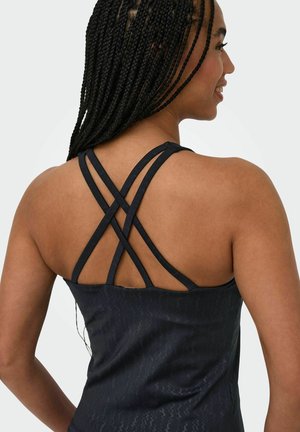 Jeune femme aux longues tresses portant un haut noir sans manches avec des fines bretelles croisées dans le dos, souriante et regardant sur le côté.