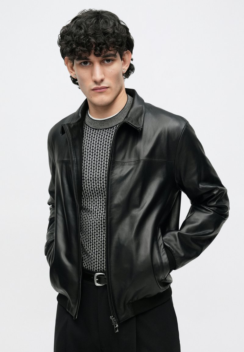 Veste en cuir noir avec une fermeture éclair et des poches latérales, associée à un pull en maille grise texturée en dessous et à un pantalon noir.