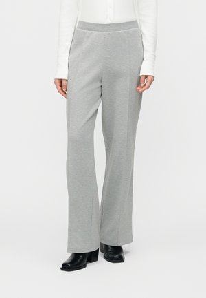 Kvinde iført lysegrå bukser med rette ben og sorte ankelstøvler med hæl, stående mod en ensfarvet lys baggrund.