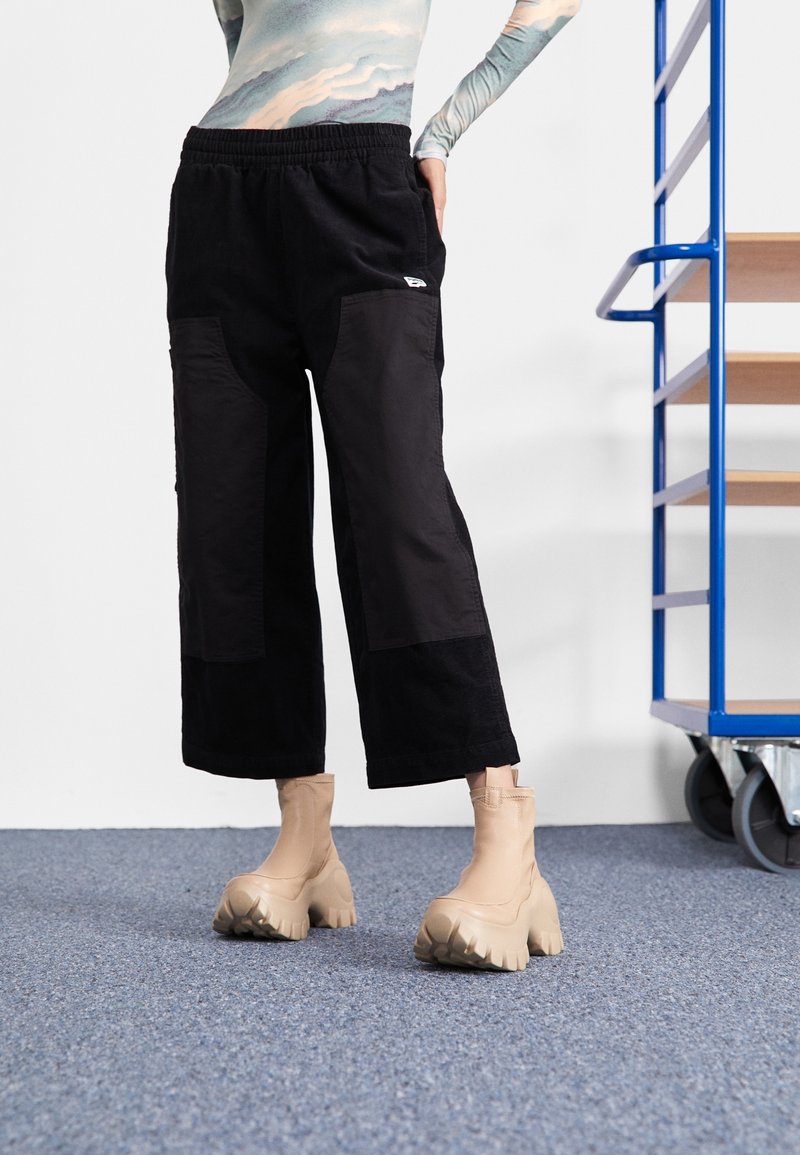 Personne portant un pantalon large noir et des bottes beige épaisses, debout sur un tapis bleu près d'une étagère à roulettes en bois et bleu.