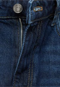Gros plan d’un jean en denim bleu foncé montrant une fermeture éclair métallique ouverte, un bouton avec gravure de la marque et des surpiqûres orange autour de la boutonnière.