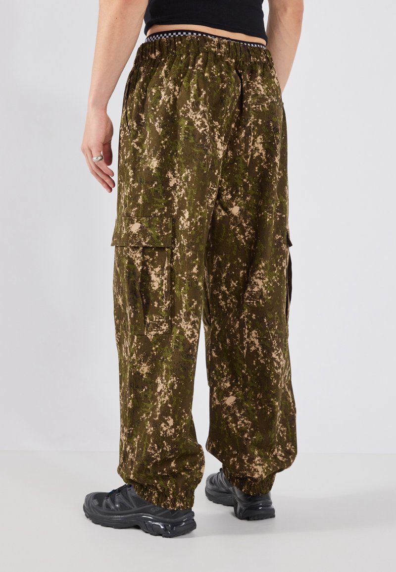 Personne portant un pantalon cargo camouflage ample et des baskets noires, debout de dos à la caméra.