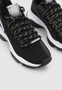 Chaussures de sport noires avec tige en tricot texturé, un design de lacets réfléchissants multicolores et une semelle intermédiaire en caoutchouc blanc avec une semelle extérieure à motifs.