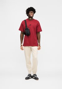 Κόκκινη βαμβακερή μπλούζα με μαύρο λογότυπο, μπεζ φόρμες και μαύρα αθλητικά παπούτσια. Το άτομο κρατά μια μικρή μαύρη τσάντα που φοριέται χιαστί.