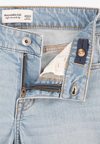 Jeans a gamba larga, di alta vita, di un blu chiaro. Presentano una zip in metallo con denti dorati, chiusura con bottone e cuciture arancioni a contrasto.