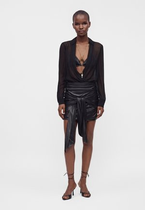 Femme portant un chemisier noir transparent à manches longues, une mini-jupe en cuir noire avec un lien à l'avant, des talons noirs avec des brides et un long collier en or.
