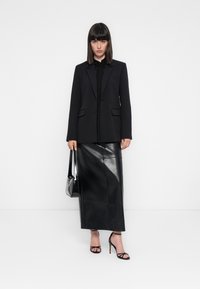 Donna in piedi contro uno sfondo bianco, indossa un blazer nero, una gonna midi in pelle, sandali con tacco alto e regge una borsa nera.