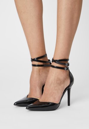 Escarpins noirs en cuir verni avec des pointes et des brides à la cheville. Les brides présentent une double boucle, et les talons font environ 10 cm de haut.
