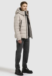 Svetlo bež pufasta jakna s kapuco, sprednjim zadrgo in črnimi žepi na zadrgo. V kombinaciji z temnimi kavbojkami in črnimi škornji z debelim podplatom.