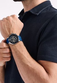 Hombre vestido con una camiseta polo azul marino ajustando un reloj cronógrafo de correa azul en la muñeca izquierda frente a un fondo blanco.