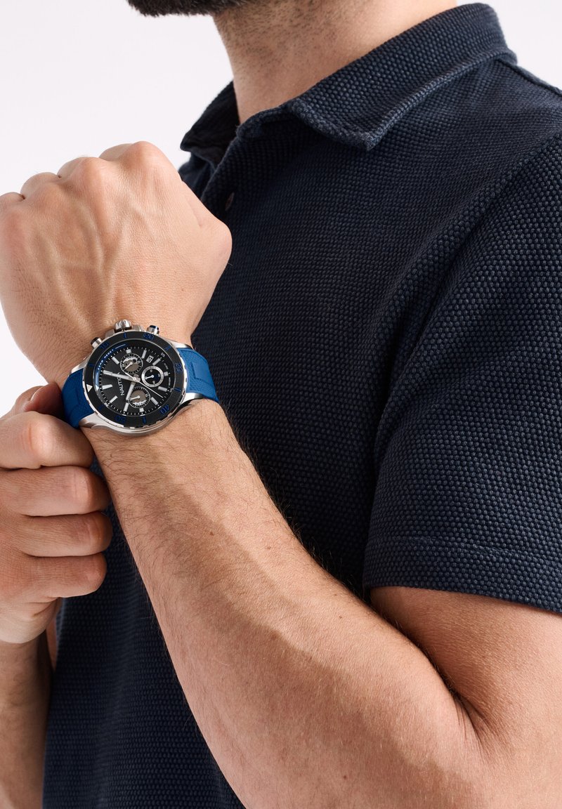 Hombre vestido con una camiseta polo azul marino ajustando un reloj cronógrafo de correa azul en la muñeca izquierda frente a un fondo blanco.