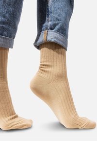 Beżowe skarpetki prążkowane, wykonane z miękkiego materiału, o długości do kostki i delikatnej fakturze. Noszone z podwiniętymi niebieskimi dżinsami.