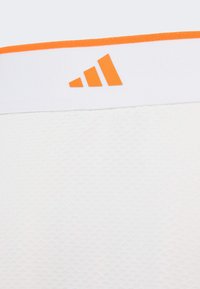 Biely športový top s textúrovaným vzorom látky a pevnou oranžovou pásikou pozdĺž výstrihu, s oranžovým logom Adidas.