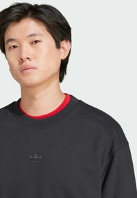 Sort sweatshirt med ribbet detaljering på skuldrene, rund hals, og en rød undertrøje der er synlig, med et lille sort logo på brystet.