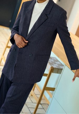 Personne portant une veste et un pantalon de costume bleu marine foncé, avec un haut blanc en dessous, debout et appuyée contre une armoire bleu clair.