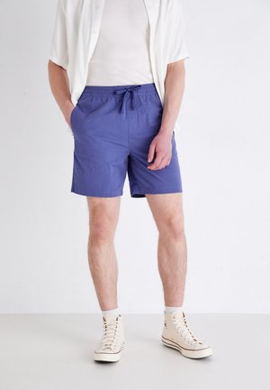 Homme portant des baskets montantes blanches, un short violet à cordon, un T-shirt blanc et une chemise à manches courtes déboutonnée, debout les mains dans les poches.
