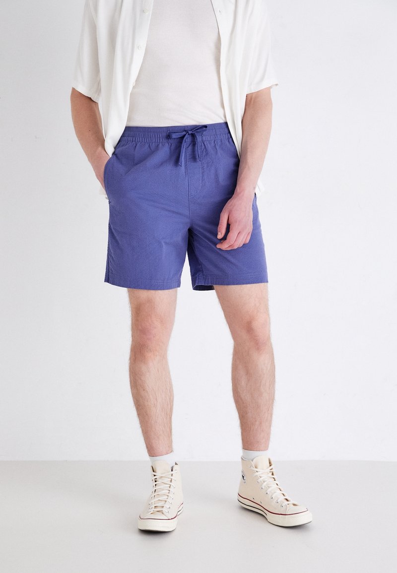 Homme portant des baskets montantes blanches, un short violet à cordon, un T-shirt blanc et une chemise à manches courtes déboutonnée, debout les mains dans les poches.