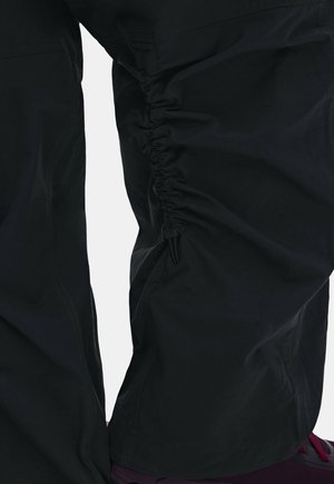 Pantalones negros con detalle elástico fruncido a lo largo del muslo externo y costuras visibles cerca de la rodilla, usados con zapatos burdeos.
