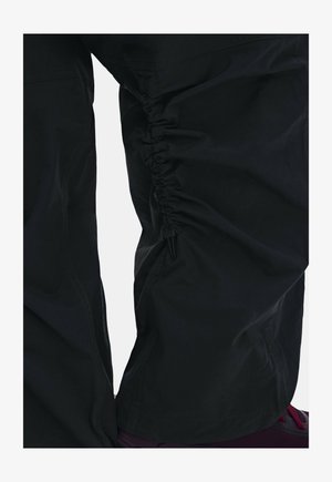 Pantalones negros con detalle elástico fruncido a lo largo del muslo externo y costuras visibles cerca de la rodilla, usados con zapatos burdeos.