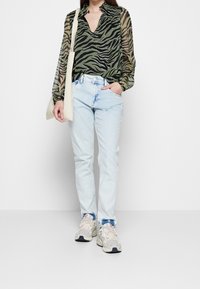 Blusa trasparente a strisce zebra verde e nera, jeans blu chiaro e sneakers voluminose. Borsa beige a tracolla.