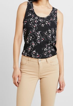 Kvinde iført ærmeløs sort top med hvidt og rødt blomsterprint og beige skræddersyede bukser, stående med armene afslappet ned langs siderne.