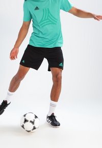 Turkusowa koszulka sportowa Adidas z czarnymi akcentami, czarne spodenki, białe skarpetki oraz czarne buty piłkarskie, ułożone obok czarno-białej piłki nożnej.