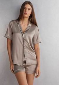 Silkig beige pyjamaset med korta ärmar och shorts, med vit piping på kragen och manchetten. Slät textur, skräddarsydd passform.
