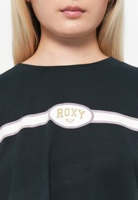 T-shirt noir avec un col rond. Présente un motif rayé blanc et rose clair sur le devant, avec un logo "ROXY" en doré.
