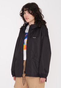 Veste légère noire avec une fermeture éclair à l'avant, des poignets élastiques et deux poches latérales. Présente un petit logo sur la poitrine gauche.