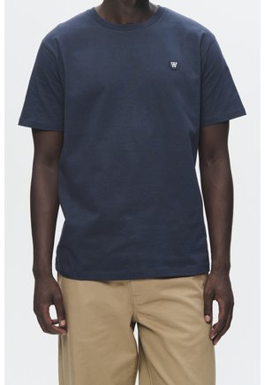 Personne à la peau foncée portant un t-shirt bleu marine uni à manches courtes avec un petit emblème blanc sur la poitrine gauche et un pantalon marron clair.