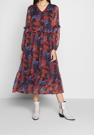 Kobieta ubrana w długą sukienkę midi z długim rękawem i dekoltem w serek, z kwiatowym wzorem w czerwone, niebieskie i czarne motywy, do tego białe botki za kostkę z czarną podeszwą.