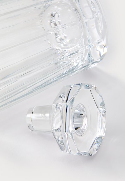 Klarer Glaskaraffe mit vertikalen Rillen, achteckigem Stopfen und glatter Textur, die Reflexionen und Lichtspiele zeigt.