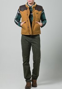 Brązowa pikowana kamizelka z beżowymi sztucznymi skórzanymi ramionami, zestawiona z zieloną i czarną koszulą w kratę oraz ciemnozielonymi spodniami. Brązowe buty na sznurowadła.