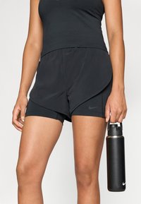 Melni sporta šorti ar iekšējo oderi, izliekta apakšmalā un Nike logo. Persona tur melnu ūdens pudeli ar sudraba vāciņu.