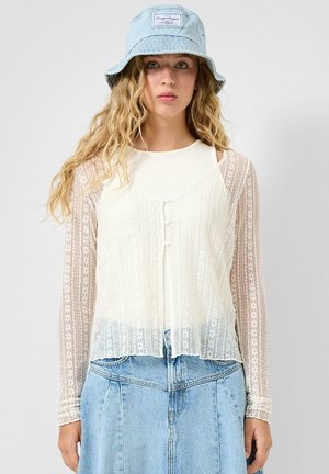 Młoda kobieta z długimi kręconymi włosami, ubrana w jasnoniebieski kapelusz typu bucket, biały koronkowy top z długim rękawem oraz jasnoniebieską spódnicę dżinsową.