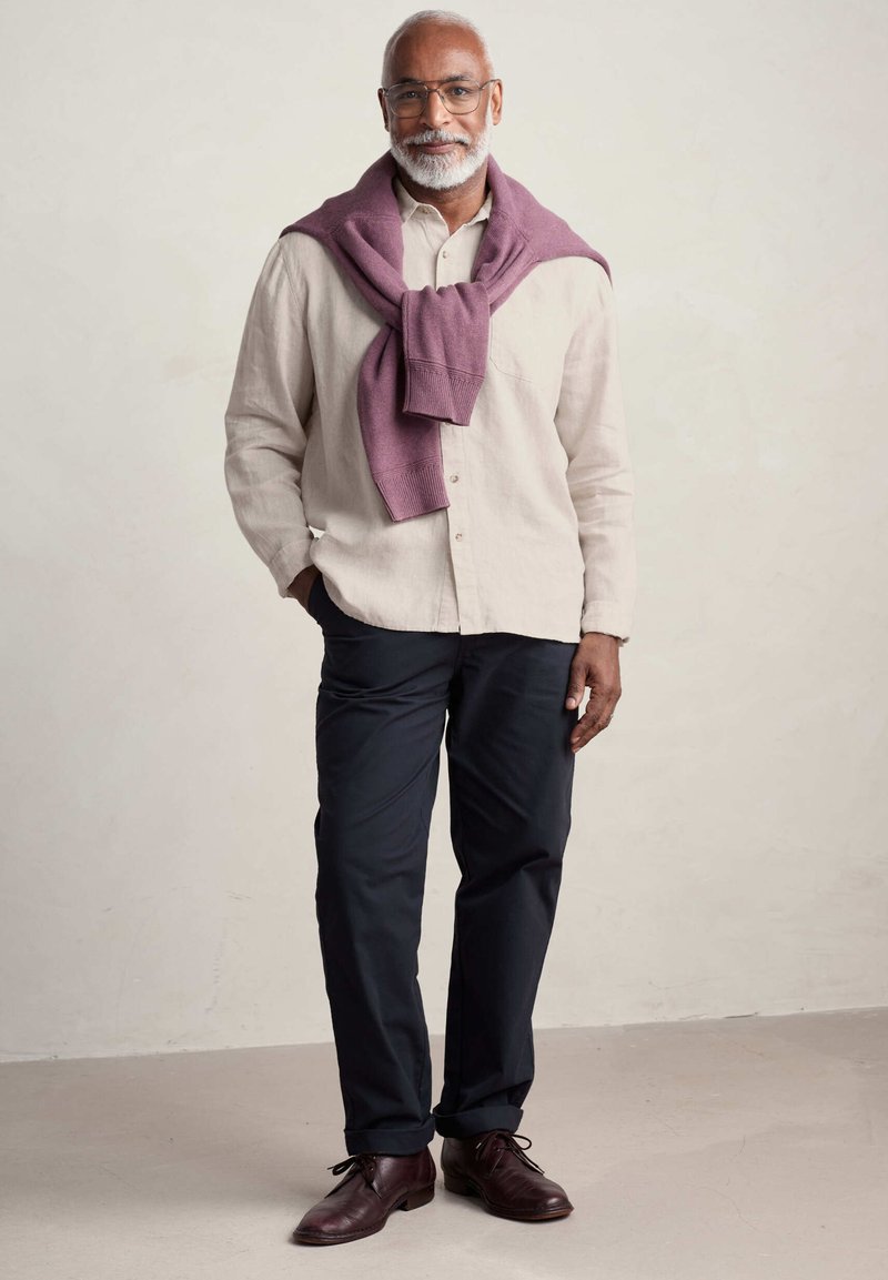 Camicia button-up di lino beige chiaro, pantaloni blu navy scuri e maglione bordeaux drappeggiato sulle spalle. Scarpe di cuoio marroni con lacci.