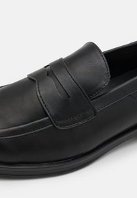 Sorte lær slip-on sko med rund tå, dekorativ stropp over vampen og subtile sømdetaljer langs sidene.