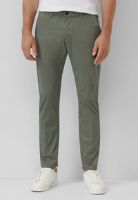 Mann in schmal geschnittenen olivgrünen Hosen und weißen Sneakers, der vor einem einfachen hellgrauen Hintergrund steht.