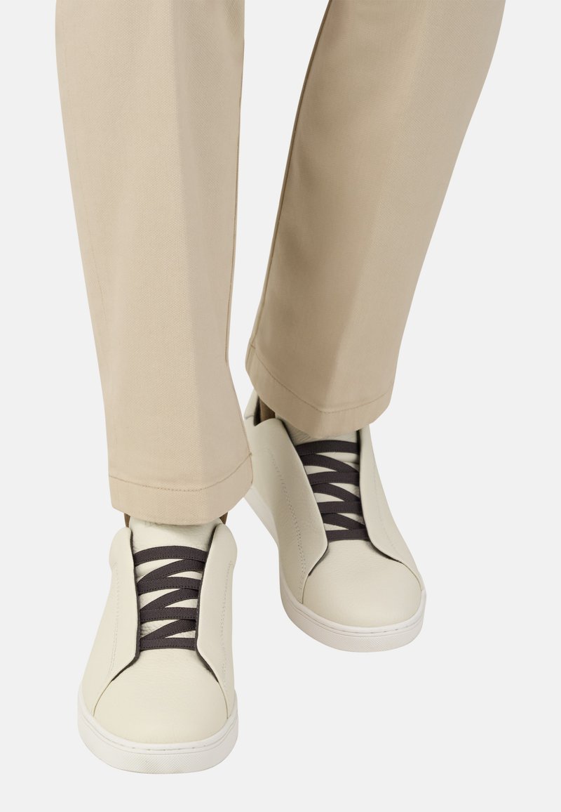 Baskets en cuir blanc avec des lacets élastiques foncés, présentant un design sans lacets et une forme minimaliste. Portées avec un pantalon droit couleur fauve.