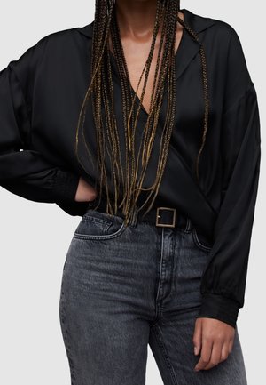 Persona con pelo largo trenzado que lleva una blusa negra de satén de manga larga metida en unos vaqueros de tiro alto de color gris oscuro con un cinturón de hebilla cuadrada.