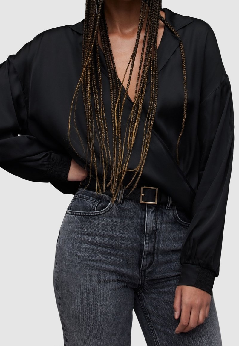 Personne aux longs cheveux tressés portant un chemisier en satin noir à manches longues, rentré dans un jean taille haute gris foncé avec une ceinture à boucle carrée.